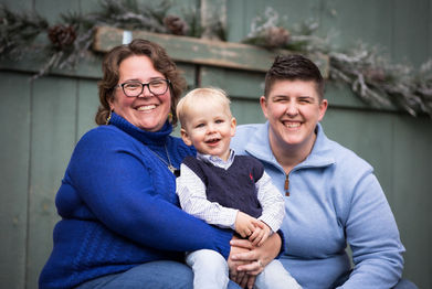Two lesbians dressed in blue sweaters sit holding their toddler son in their laps.  Green barn door with holiday greenery is in the background.  They all have big smiles.  Lesbian family photoshoot.