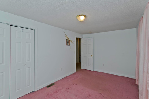 A bedroom with a slightly worn red carpet and white walls. There is a closet with white bifold doors. The bedroom door leads to a hallway.