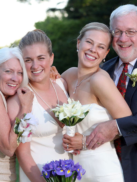 Blond brides smile as they're embraced by their parents during family portraits.