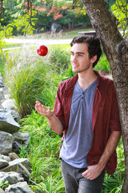 White young man with short curly dark hair leans against a tree and tosses a red apple into the air. The pastoral background includes a rock wall, tall green grasses and woods. Westford, MA