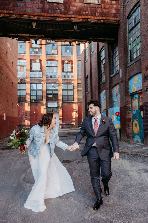 A couple holds hands as they walk outside in front of an abstract mural.