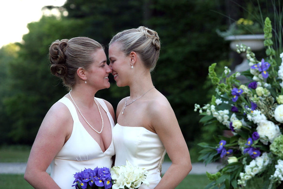 Lesbian brides smile as they are about to kiss.  Both blondes have intricate up-do hair styles adding elegance.  Both dressed in white gowns, one holds a bouquet of purple iris, the other gardenias.  Bradley Estate, Canton, MA