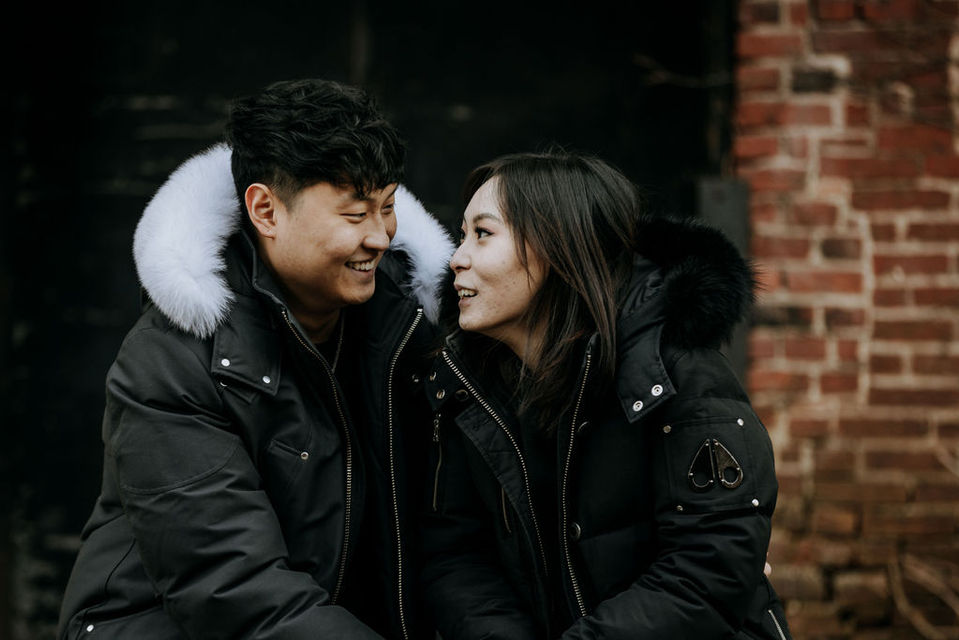 The east Asian couple sits at the edge of a condemned loading dock, a black door behind them. They look at each other smiling.