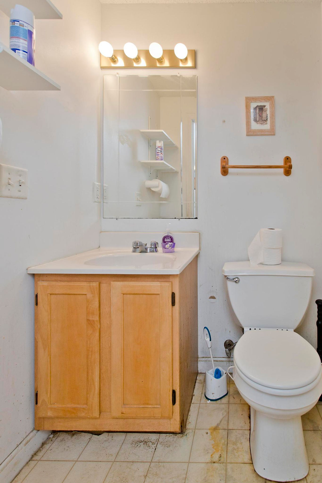 A half bathroom with wooden vanity with a white top. A mirror hangs above the vanity. There are white walls, and a white toilet.