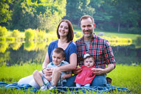 Family of four sit on grass with lake and trees behind them.  Wife leans into husband.  Husband holds infant daughter wearing red dress and bow.  Mother hugs young boy against her.  He has impish grin.  