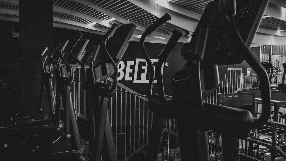 Row of empty gym ellipticals under a ribbed ceiling. A sign reads "BE FIT" in bold. The monochrome setting feels industrial.