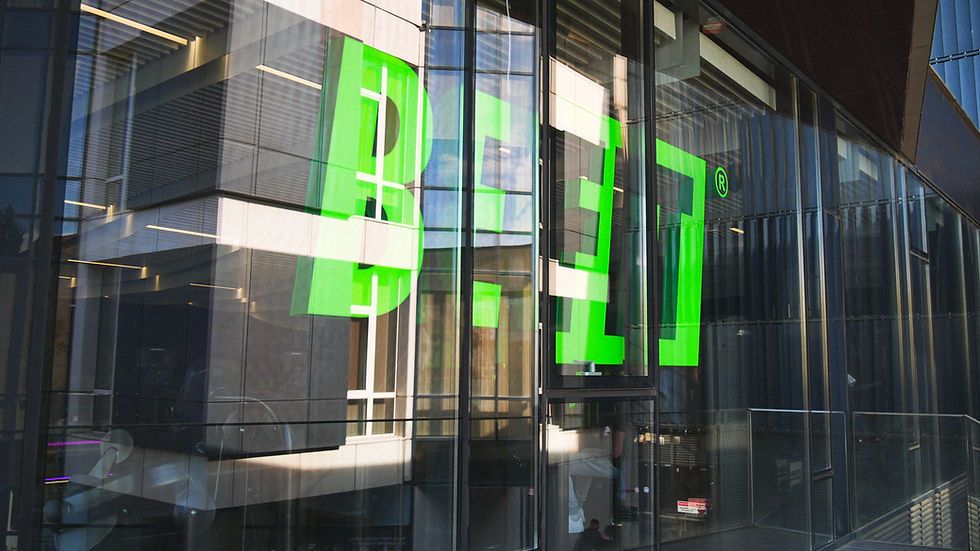 Large green letters spelling "BEAT" on a modern glass building facade, reflecting nearby structures. Bright sunlight creates dynamic patterns.