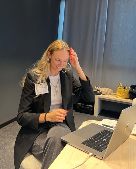 Unsere Mitarbeiterin Jil im schwarzen Blazer und mit blonden, langen Haaren sitzt mit einem Laptop an einem Schreibtisch, schaut auf ihren Laptop und lacht.