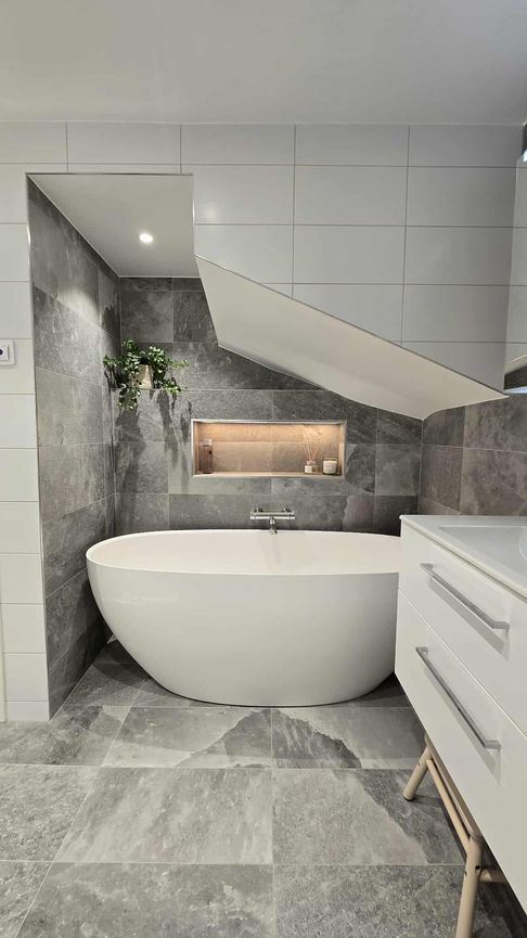 Badrum med stora grå kakelplattor, fristående badkar och infälld hylla med varm belysning. Till höger syns en vit tvättdel med tvättmaskin och torktumlare samt överskåp. Rummet har spotlights i taket och en harmonisk, hotellinspirerad känsla.