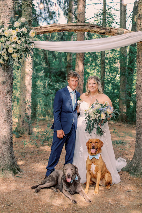a bride and a groom with their two pups in a pennsylvania forest posing underneath their luxury custom made wood and foral wedding arch