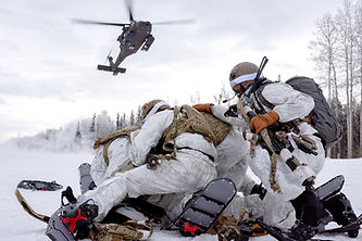 Military personelle in winter gear on a snowy mountain with a helicopter overhead