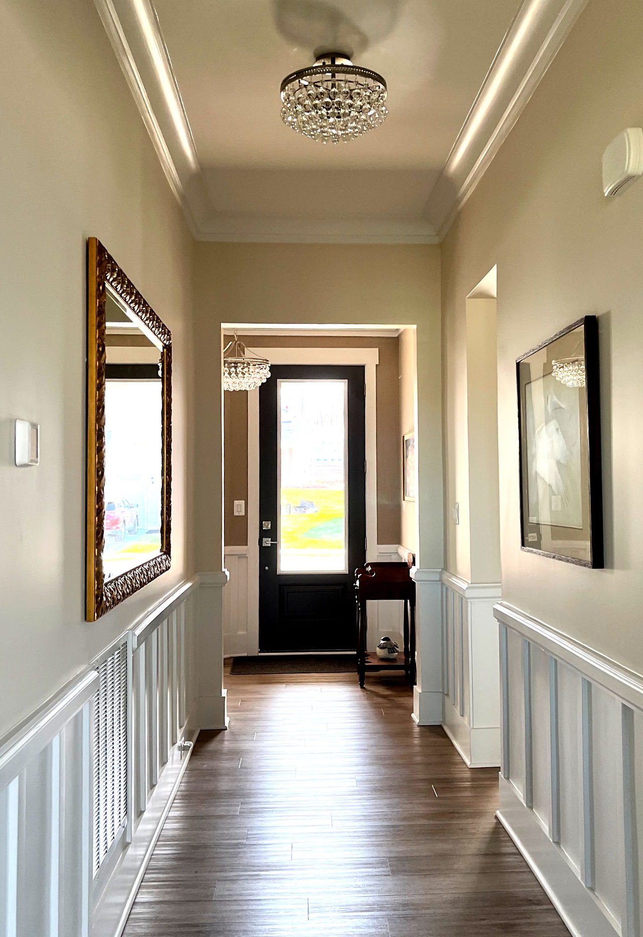 A long hallway featuring wainscaoting, large pieces of artwork, and a small crystal lighting fixture
