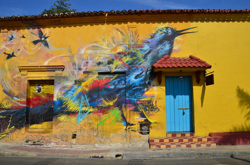 Picture of a a mustard yellow wall with the profile of a blue bird painted across it. Underneath its wings there are colorful splashes of red, green, yellow, purple, and other colors.