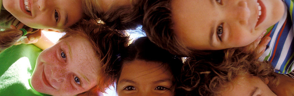 Four smiling children with diverse appearances lean in together in a circle, looking down at the camera against a bright sky.