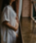 Pregnant woman in a white dress has belly exposed and is standing by a window