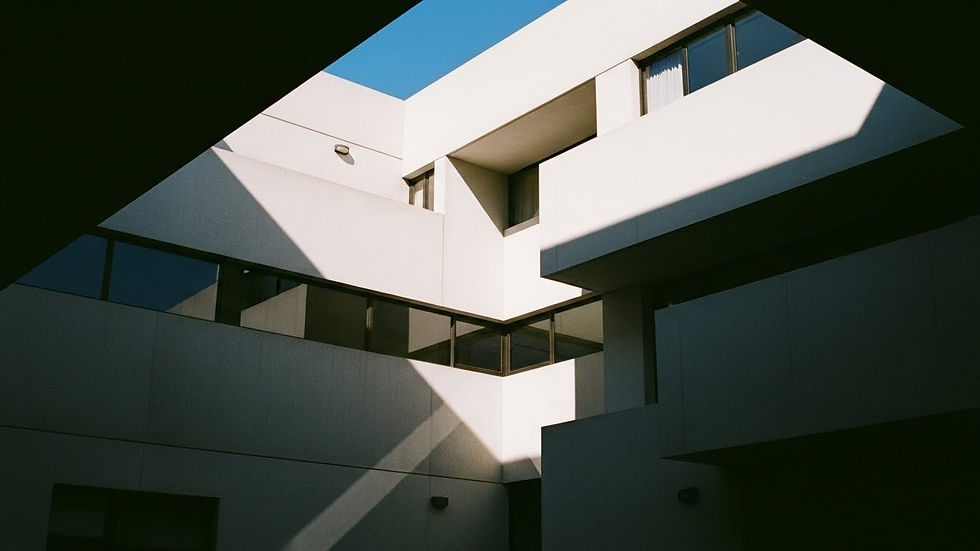 Edificio moderno de concreto blanco con sombras marcadas. Ventanas reflejan el cielo azul. Ambiente tranquilo y arquitectónico minimalista.