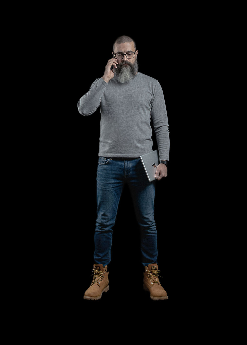 Hombre de barba hablando por teléfono, sostiene una tablet. Lleva jeans azules, suéter gris y botas marrones. Fondo negro.