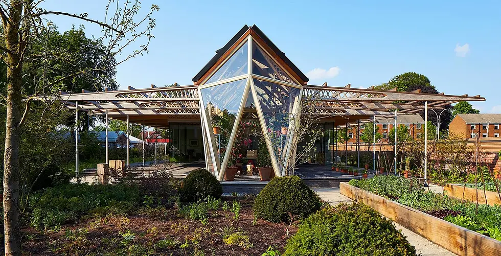 Edificio moderno con techo triangular de vidrio en un jardín. Hay plantas y árboles en primer plano. Cielo azul y casas de fondo.