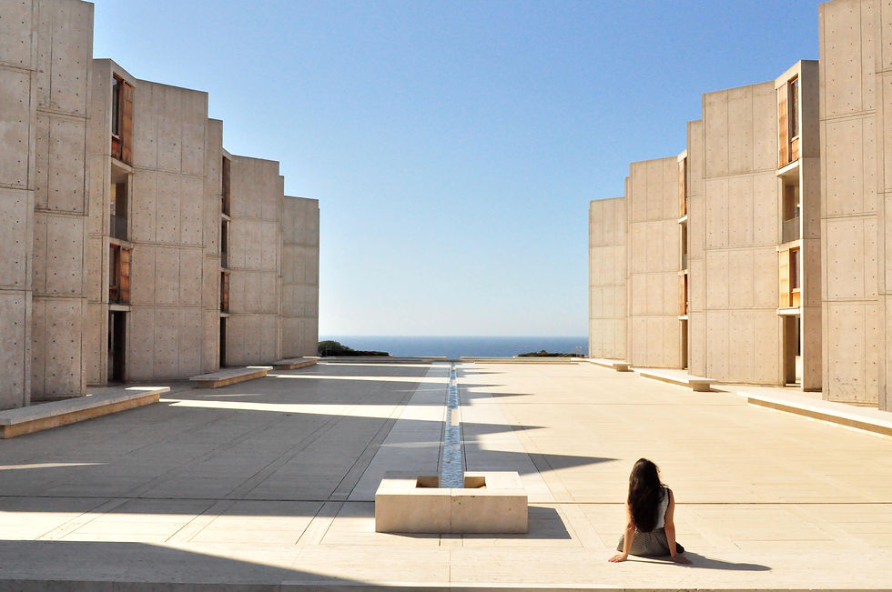 Mujer sentada en patio de un edificio de concreto, mirando al mar. Cielo despejado y luz intensa, creando sombras geométricas.