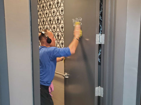 Man cleaning restroom door, removing "WCS" graffiti