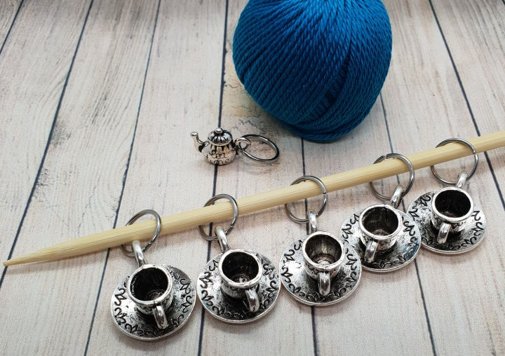 5 tea cup charms and 1 teapot charm which are silver coloured metal. Tea cup charms are on a knitting needle