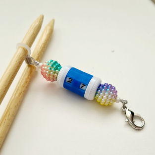 blue row counter with rainbow coloured beads at either end. A lobster clasp at one end and knitting needle hugger band at the other attached to knitting needles
