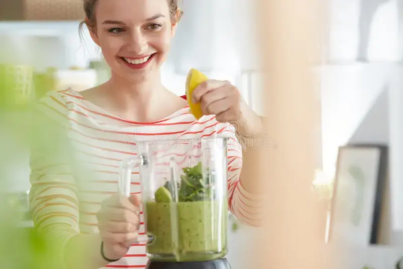 Smiling woman in a striped shirt adds lemon to a blender of green smoothie in a bright kitchen.