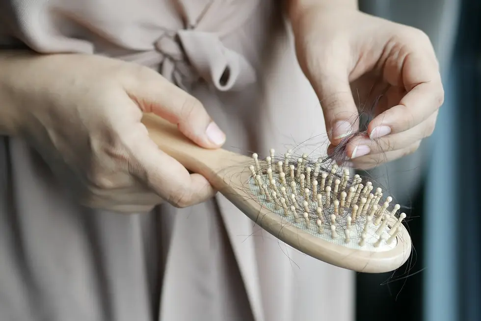 Hands pulling hair from a beige hairbrush against a soft pink top, close-up grooming scene with a few loose strands.