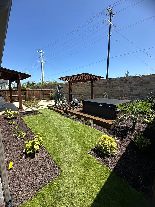 Workers installing a wooden pergola over a hot tub deck in a landscaped backyard with artificial grass and various plants.