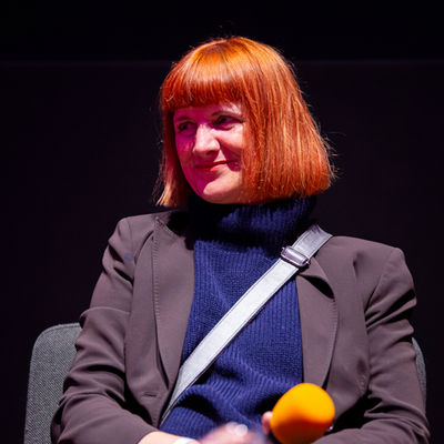 A woman with a blunt red bob haircut sitting on stage with an orange microphone resting on her lap.