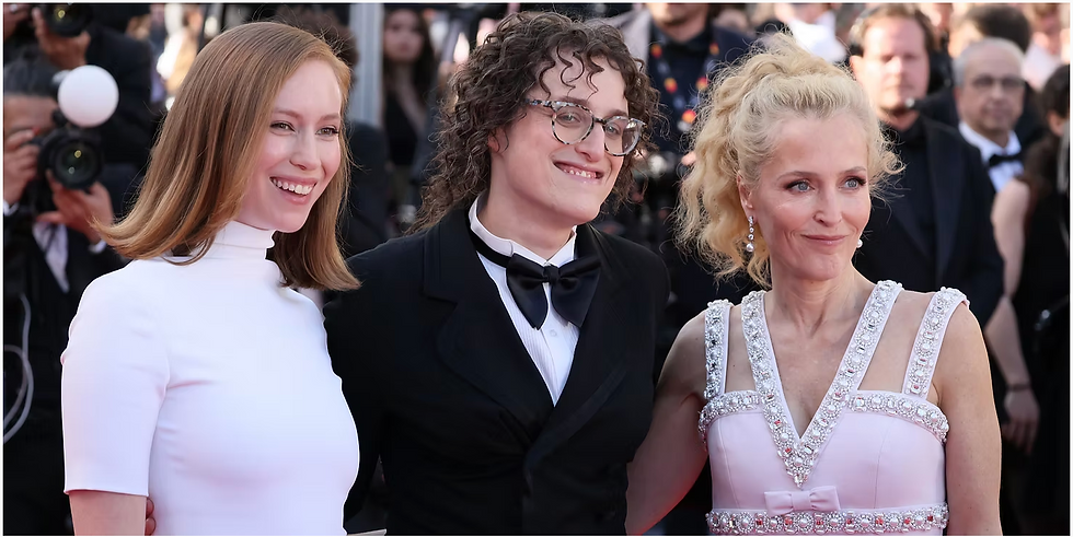 Three people pose together on a red carpet at a formal event, smiling toward photographers, dressed in elegant eveningwear and tuxedos with cameras and attendees visible in the background.