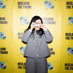 A person standing in front of a bright yellow backdrop printed with the words ‘Reclaim the Frame,’ holding a small camera up to their face as if taking a photo. They are wearing a gray suit, and the backdrop features repeated illustrated graphics alongside the text.