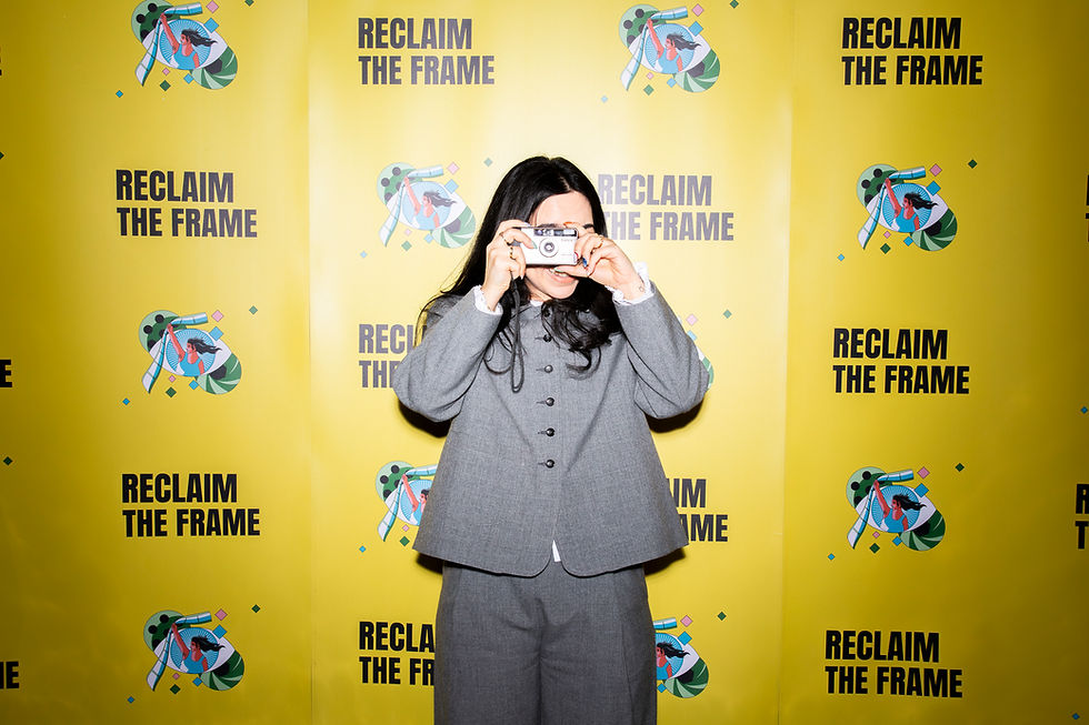 A person standing in front of a bright yellow backdrop printed with the words ‘Reclaim the Frame,’ holding a small camera up to their face as if taking a photo. They are wearing a gray suit, and the backdrop features repeated illustrated graphics alongside the text.