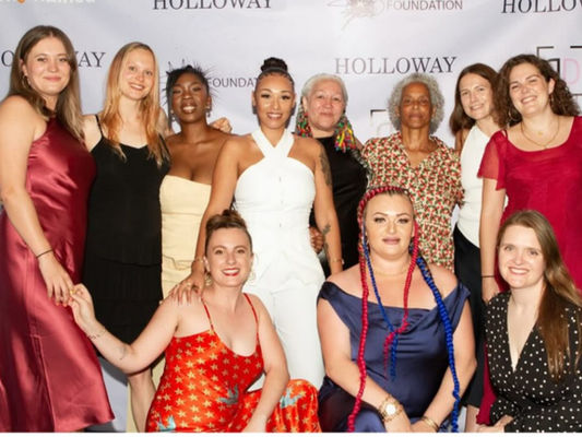 Cast and creative team in formal dresses posing on the ‘Holloway’ step-and-repeat red carpet.