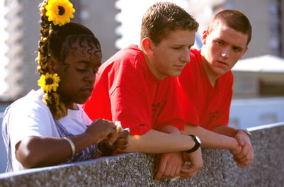 Three people sit together outdoors. A woman with braided hair and yellow flowers looks down at her hands, while two men in red T-shirts sit beside her, watching.