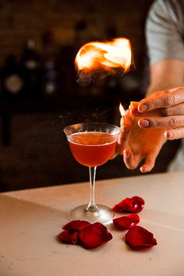 Photograph of a red drink in a clear cocktail glass- surrounded by rose petals. The glass is on fire and two hands hold a lit match.