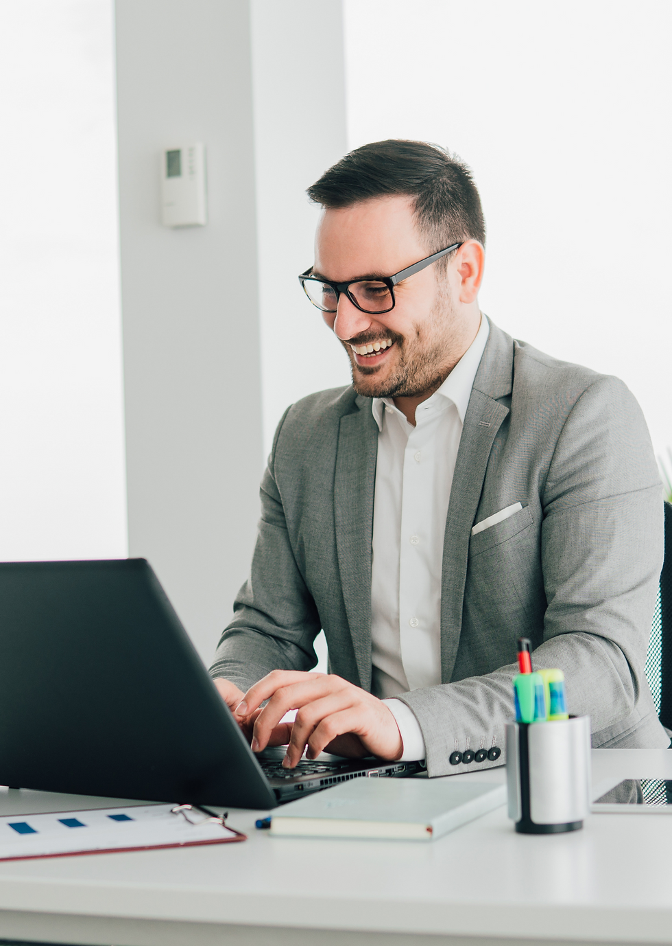 Man in a gray suit smiles while typing on a laptop at a white desk, with colorful pens in a holder. Bright, clean office setting.