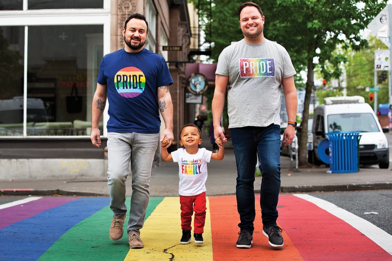 Two Dads and their son crossing a rainbow sidewalk, in celebration of Pride Month.