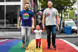 Two Dads and their son crossing a rainbow sidewalk, in celebration of Pride Month.