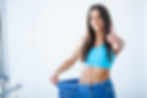 Smiling woman in blue top shows weight loss by holding out loose jeans, giving a thumbs-up. Bright, minimalist setting.