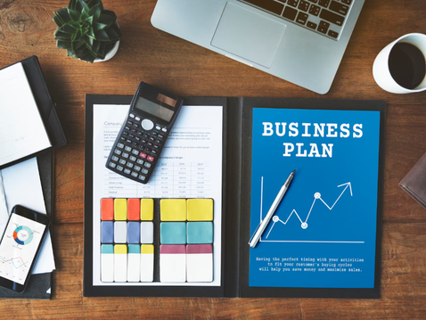 Vista superior de uma mesa de trabalho com um laptop, xícara de café, calculadora, smartphone exibindo gráficos, bloco de notas aberto e uma pasta contendo um documento intitulado 'Business Plan' ao lado de gráficos coloridos.