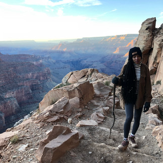 Mulher caminhando em trilha íngreme com bastão, vista panorâmica do Grand Canyon.