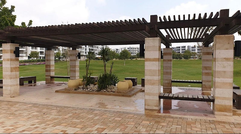 The image shows a beautiful garden in Taj City, a residential development in New Cairo, Egypt. The garden is lush and green, with a variety of flowers, trees, and shrubs. There is a central fountain in the garden, and there are several benches where people can sit and relax. The overall impression of the garden is one of peace and tranquility.
