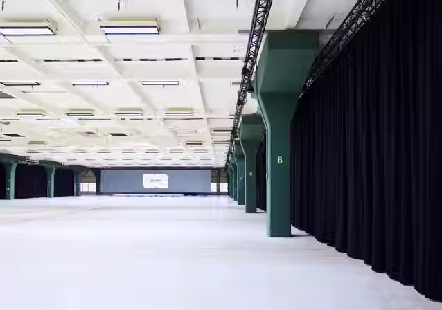 Weitläufige, helle Halle der NLND Bay 7 in Berlin mit massiven grünen Stützsäulen, hohen Decken und schwarzen Bühnenvorhängen.