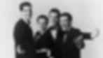 Four men in tuxedos smile and gesture broadly, posing against a plain background, conveying a joyful and lively mood.