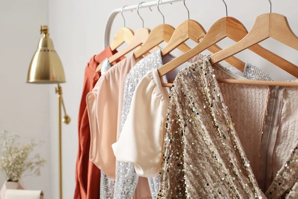 Elegant dresses in various colors, including sequins and satin, hang on wooden hangers. A stylish brass lamp and flowers in the background.