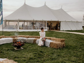 Bride in white sits on hay bales holding flowers, near a fire pit. A large white marquee tent is in the grassy background under a cloudy sky.