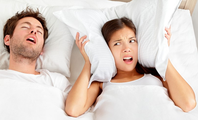Man snoring while woman covers ears with pillow, both in bed. White bedding, neutral background. Woman looks frustrated and annoyed.