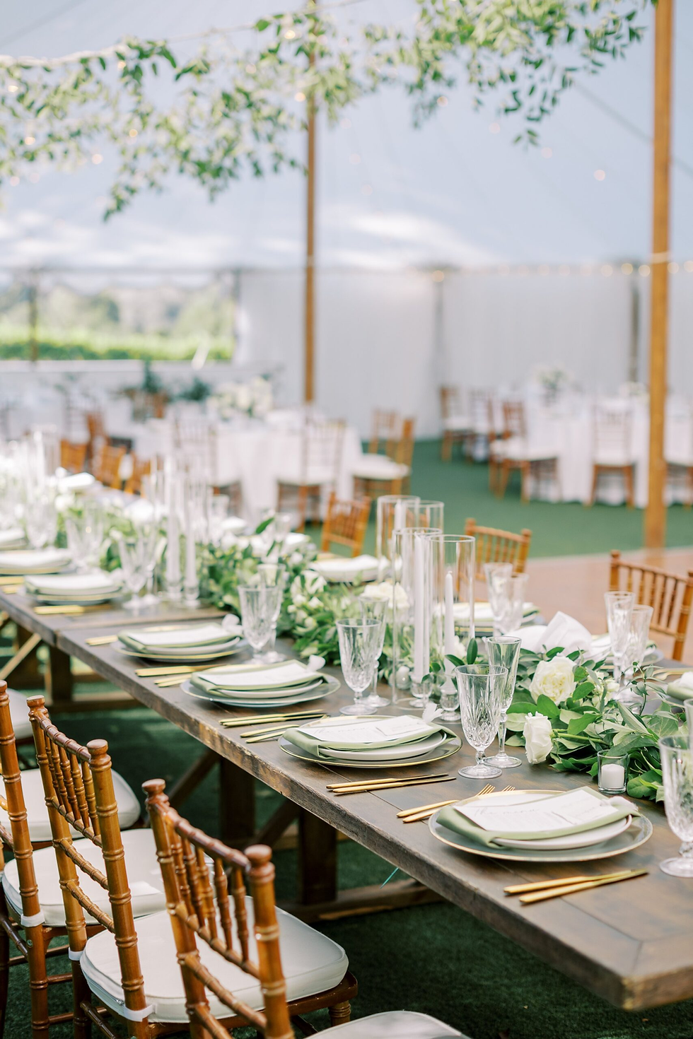 Elegant wedding table under a tent, set with green and white decor, crystal glasses, gold cutlery, and wooden chairs, creating a festive mood.