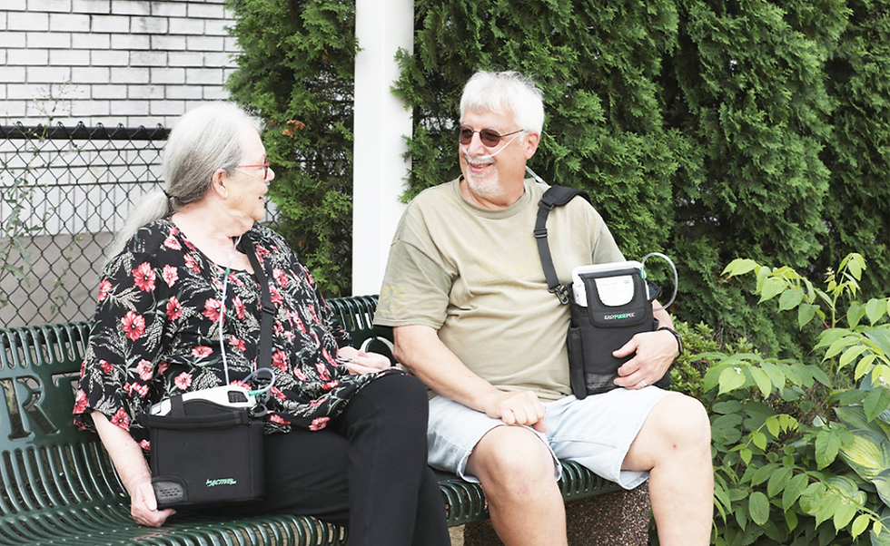 Elderly woman and man smiling on a bench, both with portable oxygen devices. Greenery and brick wall in the background. Relaxed mood.